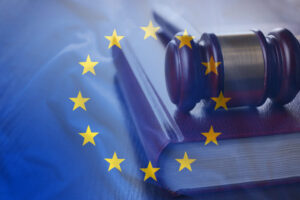 A wooden gavel rests on a closed book, overlaid with the European Union flag featuring yellow stars on a blue background. The composition suggests themes of law and governance within the EU.
