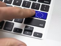 A close-up of a laptop keyboard with a unique blue key featuring the European Union flag, replacing the enter key. A finger is pressing this key. The keyboard includes black keys with white lettering and a silver casing, with part of the speaker grille visible on the right.