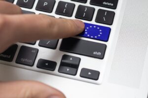 A close-up of a laptop keyboard with a unique blue key featuring the European Union flag, replacing the enter key. A finger is pressing this key. The keyboard includes black keys with white lettering and a silver casing, with part of the speaker grille visible on the right.