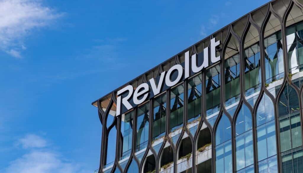 A glass office building with modern, decorative framing features the white “Revolut” logo near the roof. Blue sky and clouds are visible in the background. Green plants can be seen on some of the building’s balconies.