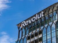 A glass office building with modern, decorative framing features the white “Revolut” logo near the roof. Blue sky and clouds are visible in the background. Green plants can be seen on some of the building’s balconies.