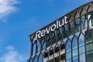 A glass office building with modern, decorative framing features the white “Revolut” logo near the roof. Blue sky and clouds are visible in the background. Green plants can be seen on some of the building’s balconies.