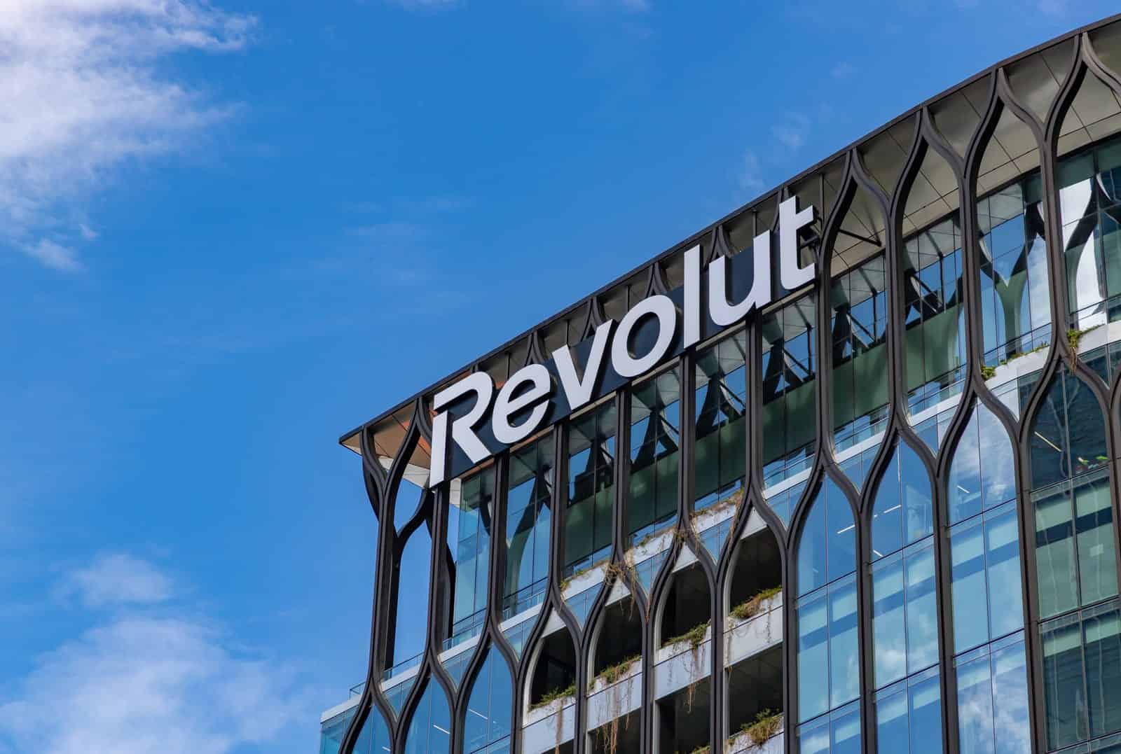 A glass office building with modern, decorative framing features the white “Revolut” logo near the roof. Blue sky and clouds are visible in the background. Green plants can be seen on some of the building’s balconies.