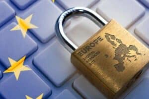 A brass padlock engraved with "EUROPE Digital Services Act" and a map of Europe sits on a white keyboard with blue keys. The EU flag, featuring yellow stars on a blue background, is overlaid on the keyboard, symbolizing digital regulation in Europe.