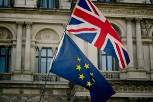 Two flags against a stone building: The Union Jack on top, featuring red, white, and blue, and the European Union flag beneath, blue with a circle of yellow stars. The building has ornate carvings and sculptures visible in the background.