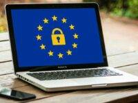 A laptop on a wooden table displays a blue screen with a yellow padlock surrounded by twelve stars, symbolizing the European Union. A smartphone lies next to the laptop. The setting appears to be outdoors, with blurred greenery in the background.