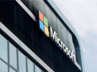 The image shows a modern building with the Microsoft logo. The logo consists of four colored squares—red, green, blue, and yellow—forming a larger square beside the word "Microsoft" in white letters. The facade features glass windows and dark vertical panels.