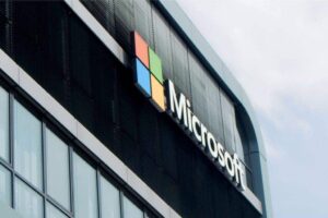 The image shows a modern building with the Microsoft logo. The logo consists of four colored squares—red, green, blue, and yellow—forming a larger square beside the word "Microsoft" in white letters. The facade features glass windows and dark vertical panels.