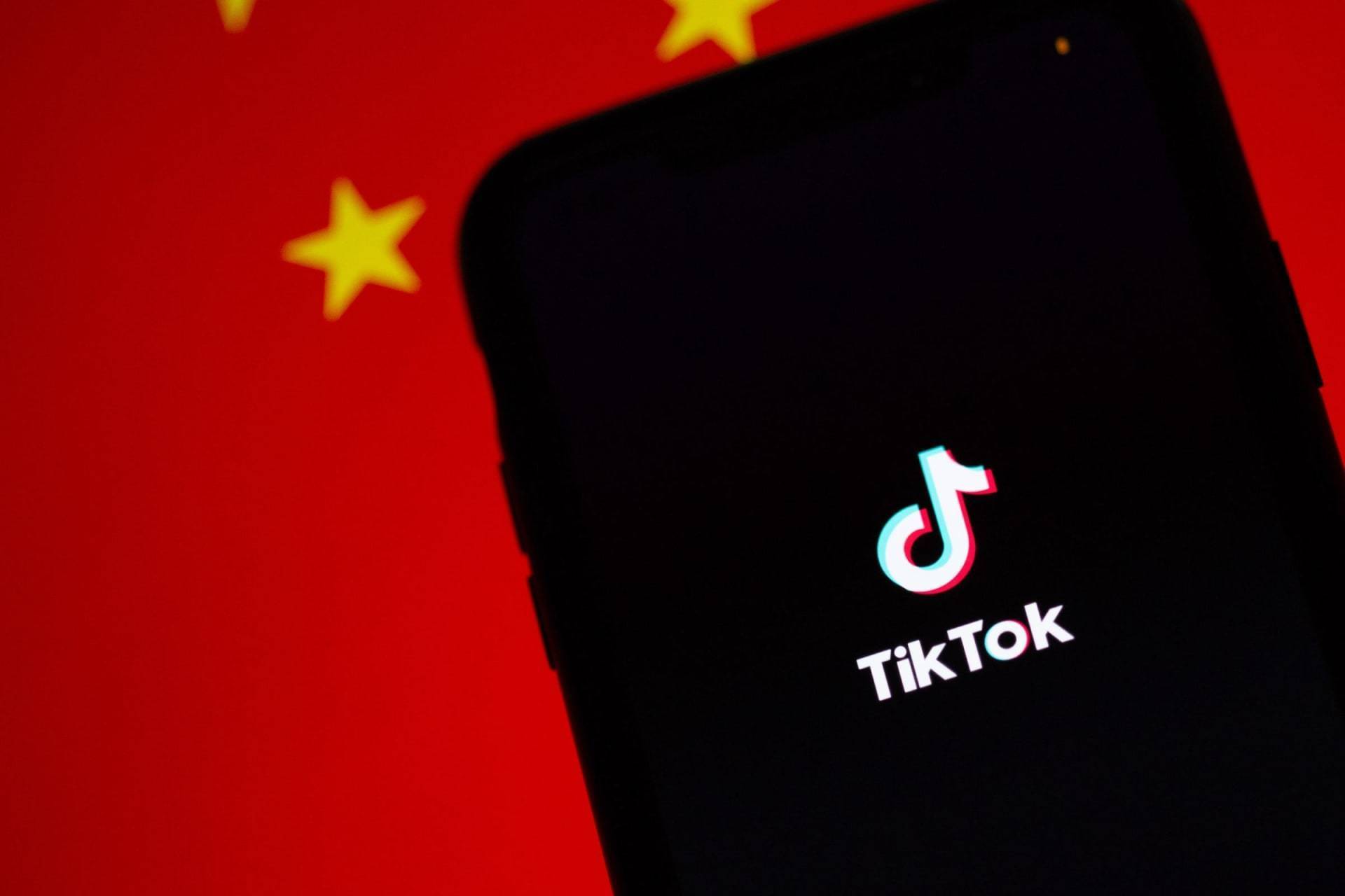 Smartphone displaying the TikTok logo on its screen, set against a background featuring China's red flag adorned with yellow stars. The image juxtaposes the digital app with the national symbol.