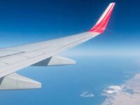 Airplane wing with a red tip against a clear blue sky. Below, there is an expanse of ocean meeting a sandy coastline, with some white clouds near the coast. The horizon divides the blues of the sky and sea, creating a serene aerial scene.