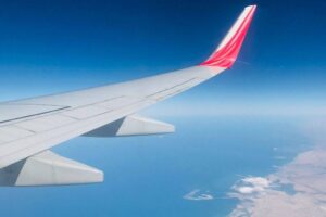 Airplane wing with a red tip against a clear blue sky. Below, there is an expanse of ocean meeting a sandy coastline, with some white clouds near the coast. The horizon divides the blues of the sky and sea, creating a serene aerial scene.