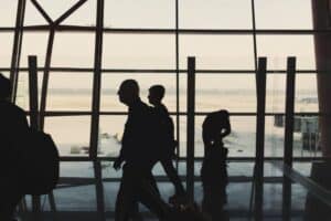 Silhouetted figures walk or stand in an airport terminal, with large windows showing an empty runway background. One person leans over luggage, while others move forward. The scene is backlit by daylight, creating distinct outlines against the glass.