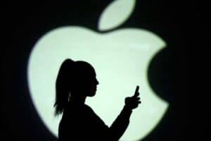Silhouette of a person holding a smartphone against a large, illuminated Apple logo backdrop. The person is seen in profile, with their hair in a ponytail, creating a stark contrast with the bright logo. The scene is minimalistic, with a focus on technology and the brand.