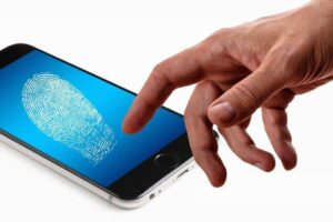 A person's hand extends toward a smartphone lying screen-up on a white surface. The phone displays a large white fingerprint on a blue background, symbolizing biometric security or fingerprint scanning. The image suggests unlocking the phone using a fingerprint.