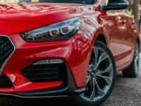 Close-up of the front right side of a shiny red car in a natural setting. The focus is on the sleek design featuring a modern headlight, honeycomb grille, and stylish alloy wheel. Tree reflections are visible on the hood, enhancing the polished look of the vehicle.