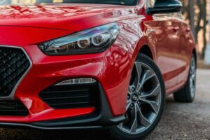 Close-up of the front right side of a shiny red car in a natural setting. The focus is on the sleek design featuring a modern headlight, honeycomb grille, and stylish alloy wheel. Tree reflections are visible on the hood, enhancing the polished look of the vehicle.