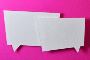 Two overlapping white speech bubble cutouts on a vibrant pink background. The cutouts have a textured appearance, and both point towards the bottom left. The bold contrast between the white speech bubbles and pink background creates a striking visual effect.