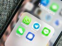 Close-up of a smartphone screen displaying a folder labeled "Messages" with five messaging app icons: WhatsApp, Telegram, WeChat, LINE, and Signal. The background shows a blurred view of more app icons. The phone edge is visible on a textured surface.