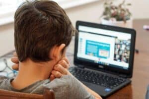 A child with short brown hair is sitting at a wooden table, facing a laptop screen. The screen displays a virtual classroom or online learning platform. The child rests their chin on their hands. A potted plant is in the background on the table.