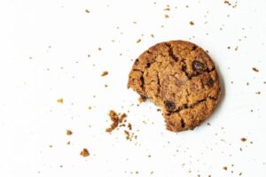 A chocolate chip cookie with a bite taken out rests on a white background. Crumbs are scattered around, adding texture to the scene. The cookie is golden brown with visible chocolate chips, and its rough, crumbly texture is evident.