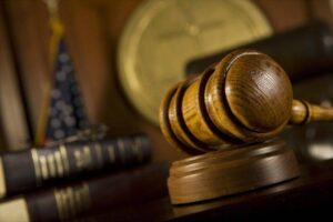 Close-up of a wooden judge's gavel resting on its base. Behind the gavel, there's a blurred American flag and stacks of legal books against a dark wooden background. The lighting gives the scene a formal and authoritative ambiance, suggesting a courtroom or legal setting.