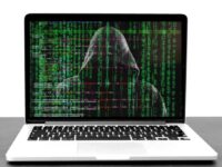 A laptop displays an image of a hooded figure with green binary code cascading down the screen, resembling hacking or cybersecurity themes. The background is a dark digital matrix with lines of numbers and symbols. The laptop sits on a plain surface.