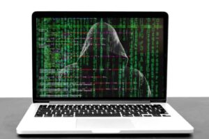 A laptop displays an image of a hooded figure with green binary code cascading down the screen, resembling hacking or cybersecurity themes. The background is a dark digital matrix with lines of numbers and symbols. The laptop sits on a plain surface.
