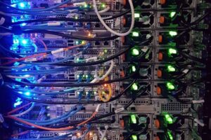 Close-up of a server rack filled with several servers. Numerous colored cables—blue, white, and black—are connected to the servers. Green LED lights indicate active connections, while orange buttons are visible on the hardware. The servers have metallic casings with ventilation panels.