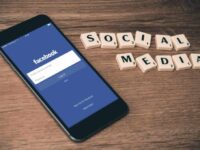 A smartphone displaying the Facebook login screen is placed on a wooden table. Next to the phone, Scrabble tiles spell out "SOCIAL MEDIA." The scene suggests a focus on social media platforms and online connectivity.