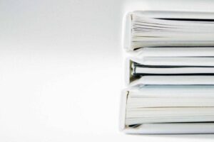 Three closed white binders are stacked horizontally against a plain white background. The binders are aligned neatly, with their spines facing right. Their pages are slightly visible and appear to be filled with documents.