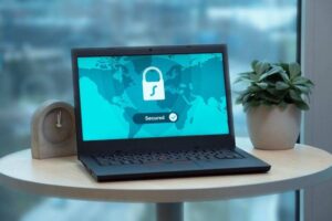 A laptop displaying a digital lock icon with the word "Secured" and a checkmark on the screen is placed on a round wooden table. The screen shows a map of the world in the background. A small gray clock and a potted plant are also on the table, with a window view behind them.