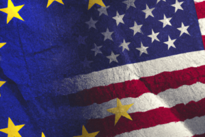 A digitally manipulated image blending the EU and US flags. The stars from the EU flag overlay the blue field of stars from the US flag, while the red and white stripes of the US flag merge into the blue background of the EU flag, creating a textured, intertwined appearance.