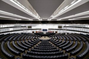 Spacious, empty auditorium with semicircular seating facing a central podium. Multiple rows of black chairs and desks. High, illuminated ceiling with white panels. Flags visible behind the podium, indicating an international setting. Large digital screen centered above.