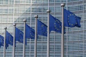 A row of European Union flags, featuring yellow stars on a blue background, fluttering on flagpoles. They are set against a modern building facade with a grid of windows. The perspective captures the flags in diminishing order, creating a sense of depth and unity.