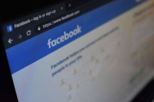 Close-up of a computer screen displaying the Facebook login page. The browser tab shows "Facebook - log in or sign up" with a secure connection icon and URL. The Facebook logo is prominently placed, and blurred content on the page is visible below.