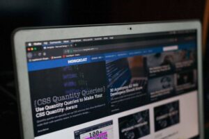 A laptop screen displays a website with articles on technology and design. The visible article titled "CSS Quantity Queries" is open, discussing CSS features. The website's header includes navigation links, and several thumbnail images with article titles are visible below.