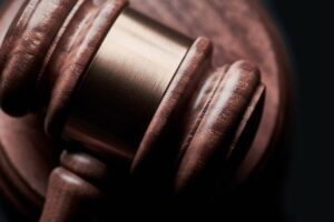 Close-up of a wooden gavel resting on a sound block, focusing on the intricate texture and grain of the polished wood. The metal band around the gavel's head is slightly blurred, emphasizing the craftsmanship and legal symbolism. Background is dark, creating contrast.