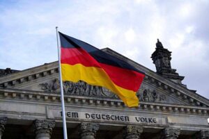German flag waves in front of the Reichstag building in Berlin. The flag displays horizontal stripes of black, red, and gold. The building's facade shows intricate sculptures and the inscription "DEM DEUTSCHEN VOLKE." The sky is cloudy.