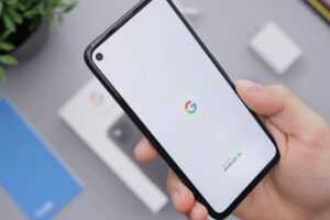 Close-up of a hand holding a smartphone with a white screen displaying the Google logo and "powered by Android" text. In the background, there's a phone box, a plant, a blue booklet, and a charger on a gray surface.