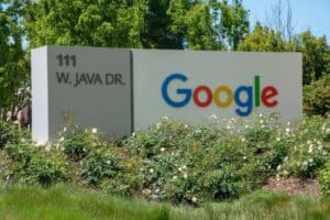 A sign displaying "111 W. Java Dr." and the Google logo in colorful letters is surrounded by greenery and white flowers. The background features trees and a clear blue sky, indicating a sunny day.
