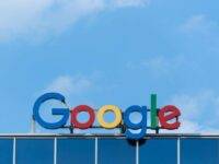 The image shows the Google logo on the top of a building against a clear blue sky. The logo features the word "Google" in colorful letters: blue, red, yellow, and green. The building's facade is made of glass panels, reflecting the sky.