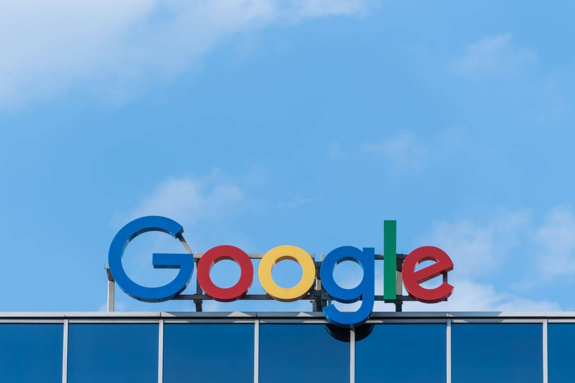 The image shows the Google logo on the top of a building against a clear blue sky. The logo features the word "Google" in colorful letters: blue, red, yellow, and green. The building's facade is made of glass panels, reflecting the sky.