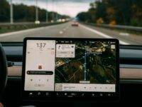 The image shows a Tesla car dashboard with a large touchscreen display. It shows navigation on a highway, speed of 137 km/h, and a map with directions to a zoo. Music is playing. The view through the windshield shows a road with trees on the sides and a car ahead.