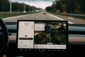 The image shows a Tesla car dashboard with a large touchscreen display. It shows navigation on a highway, speed of 137 km/h, and a map with directions to a zoo. Music is playing. The view through the windshield shows a road with trees on the sides and a car ahead.