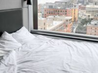 Unmade bed with white sheets and pillows next to a large window. Outside, an urban cityscape with tall buildings and a variety of architectural styles is visible. The scene is bright, suggesting daytime, and the view captures a mix of modern and older structures.