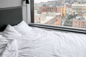 Unmade bed with white sheets and pillows next to a large window. Outside, an urban cityscape with tall buildings and a variety of architectural styles is visible. The scene is bright, suggesting daytime, and the view captures a mix of modern and older structures.