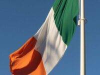 The image shows the Irish flag waving against a clear blue sky. The flag features three vertical stripes: green on the left, white in the center, and orange on the right. It is attached to a white flagpole, and the flag is gently billowing in the breeze. The image shows the Irish flag waving against a clear blue sky. The flag features three vertical stripes: green on the left, white in the center, and orange on the right. It is attached to a white flagpole, and the flag is gently billowing in the breeze.