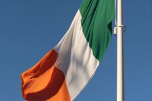 The image shows the Irish flag waving against a clear blue sky. The flag features three vertical stripes: green on the left, white in the center, and orange on the right. It is attached to a white flagpole, and the flag is gently billowing in the breeze.