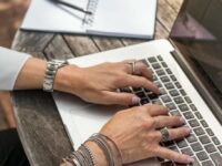 A person with multiple rings and bracelets types on a laptop placed on a weathered wooden table. A spiral-bound notebook with an open page and a silver pen is in the background, creating an outdoor work setting. The person's fingernails are painted light lavender.