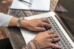 A person with multiple rings and bracelets types on a laptop placed on a weathered wooden table. A spiral-bound notebook with an open page and a silver pen is in the background, creating an outdoor work setting. The person's fingernails are painted light lavender.